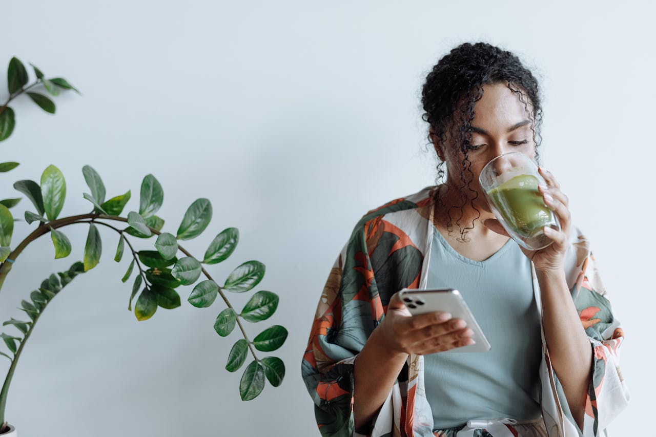 A woman sips matcha tea as she checks her phone, surrounded by indoor plants.